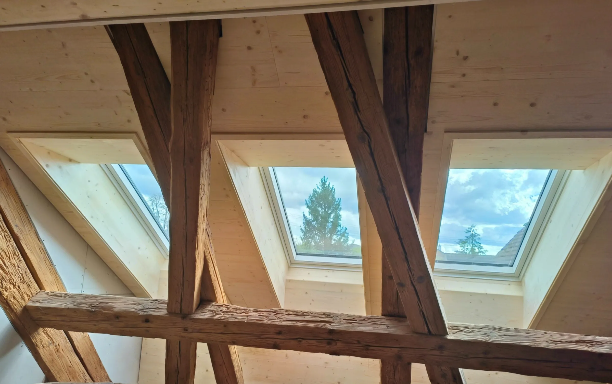 Drei Dachfenster mit hellen Futtern in einer Decke aus Dreischichtplatten hinter alten Holzbalken, Kaiseraugst
