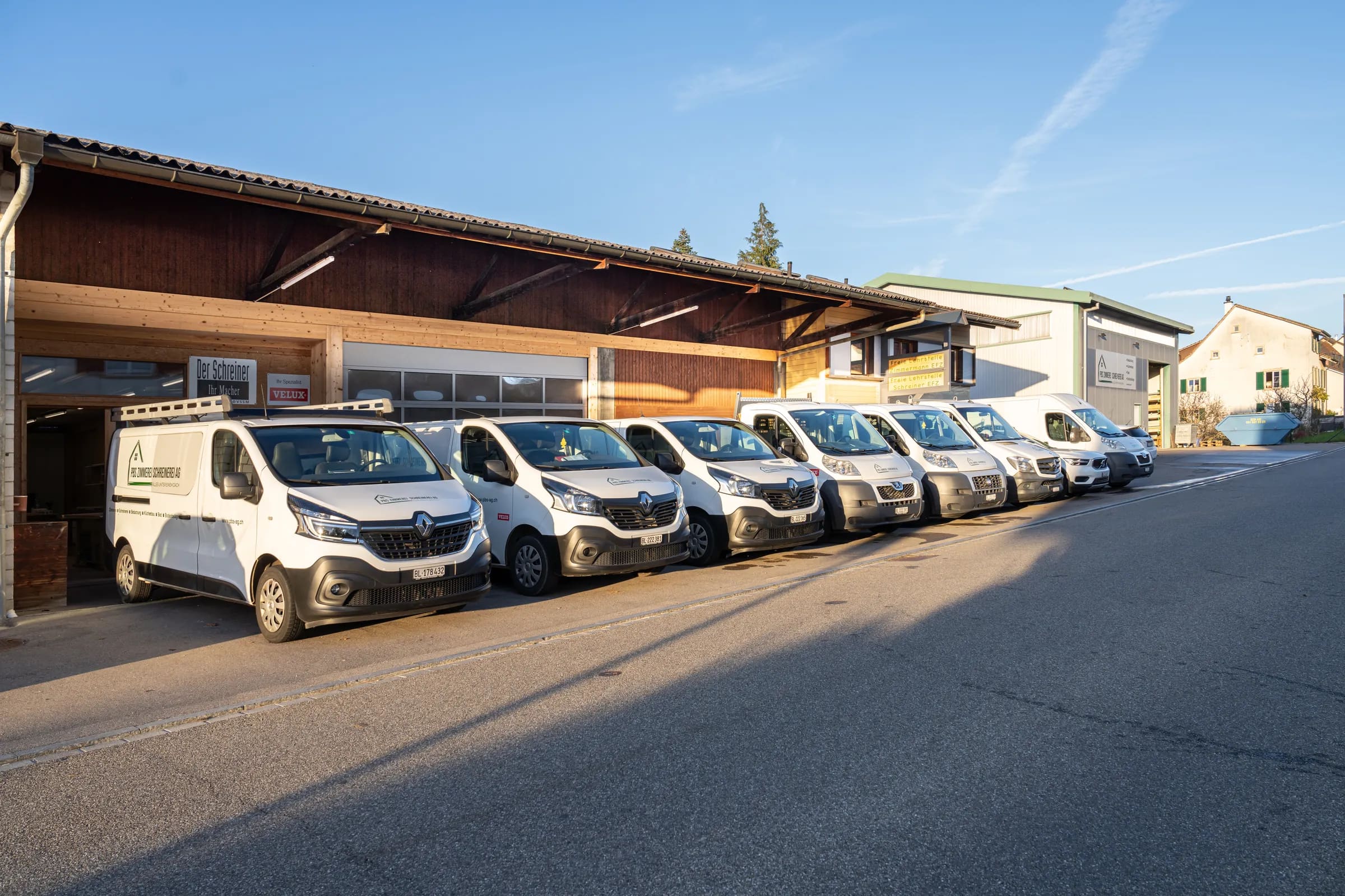 Sieben weisse Firmentransporter mit der Aufschrift «Alles unter einem Dach» vor der Werkhalle mit Holzfassade