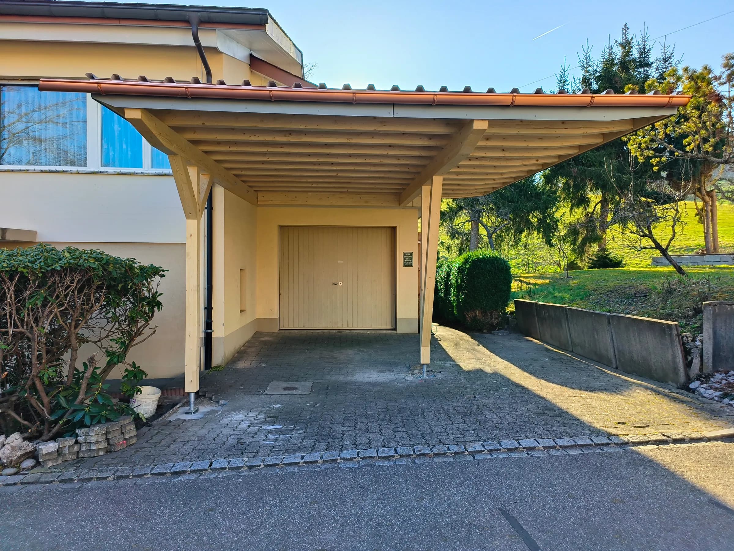 Holz-Carport mit Ziegeldach vor einem Garagentor in Niederdorf, gepflästerter Vorplatz