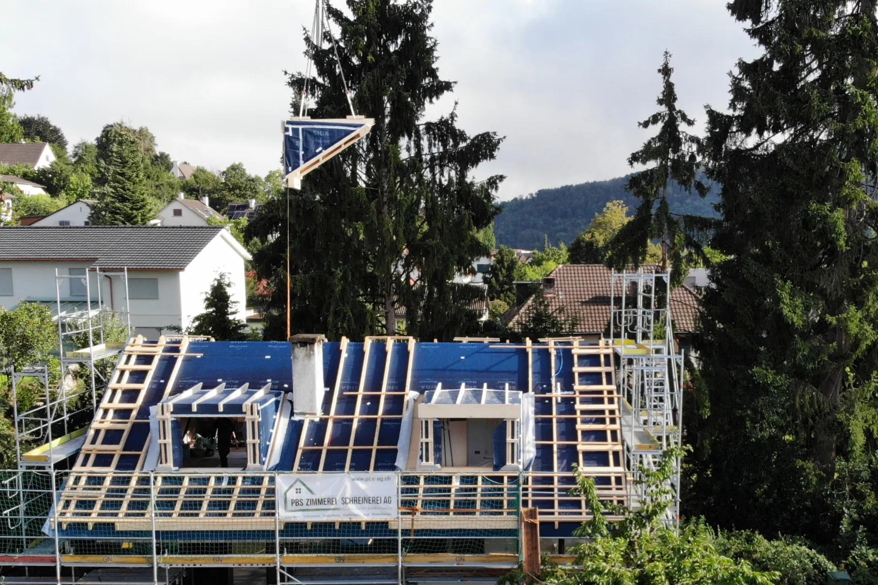 Kran hebt ein Bauteil auf das eingerüstete Haus, neue Lukarnen im Rohbau auf blauer Unterdachbahn