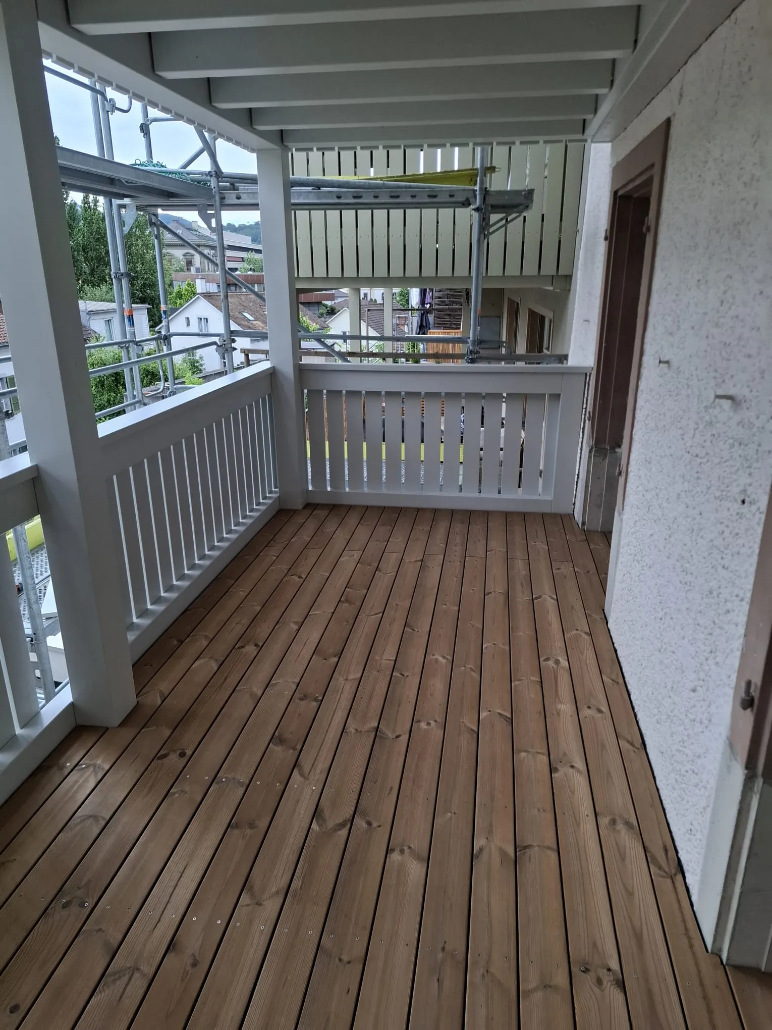 Laube mit Holzboden und weissem Geländer, dahinter das Gerüst