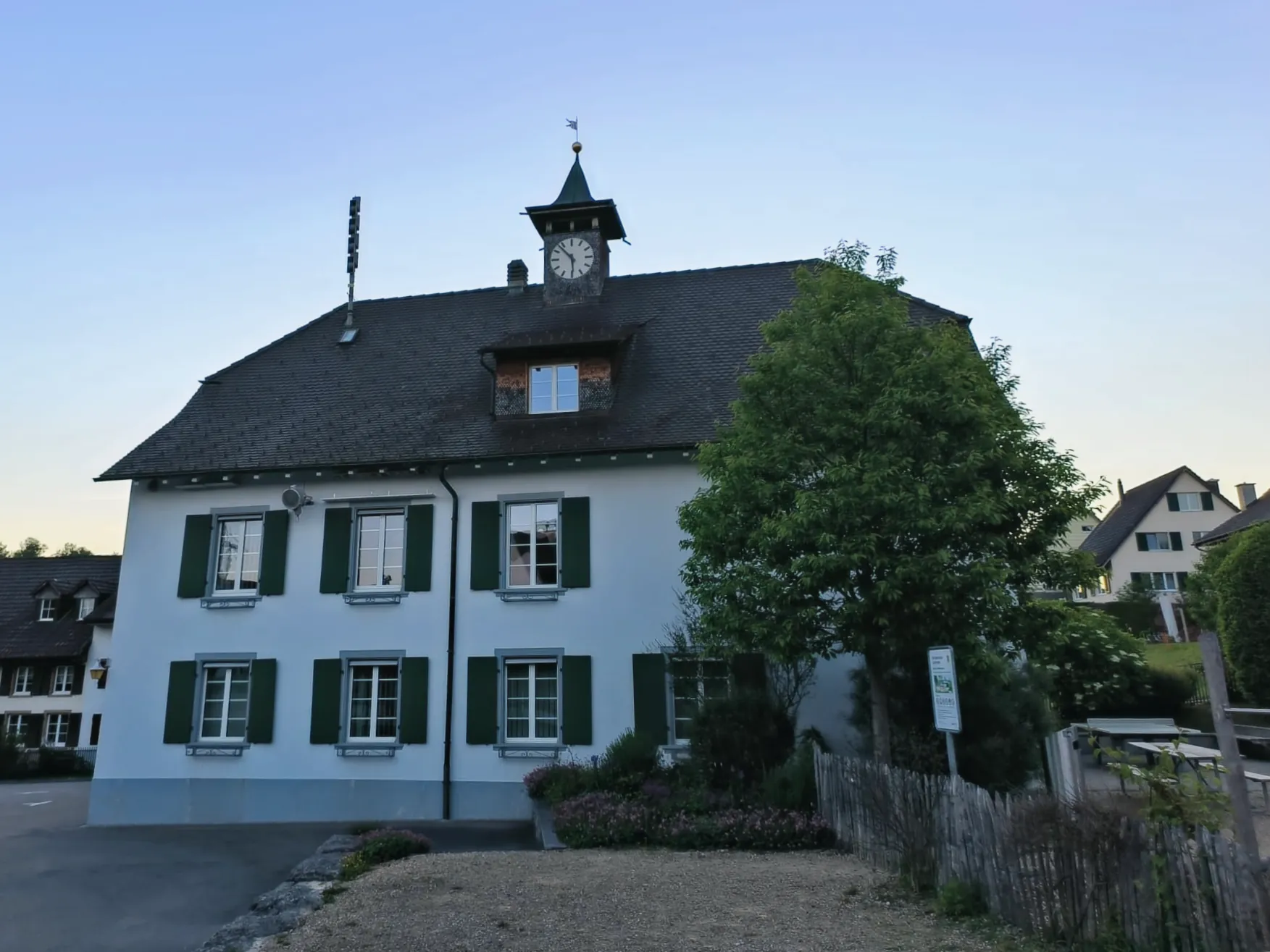 Gemeindehaus Lupsingen im Abendlicht: weisses Gebäude mit grünen Fensterläden und Uhrtürmchen