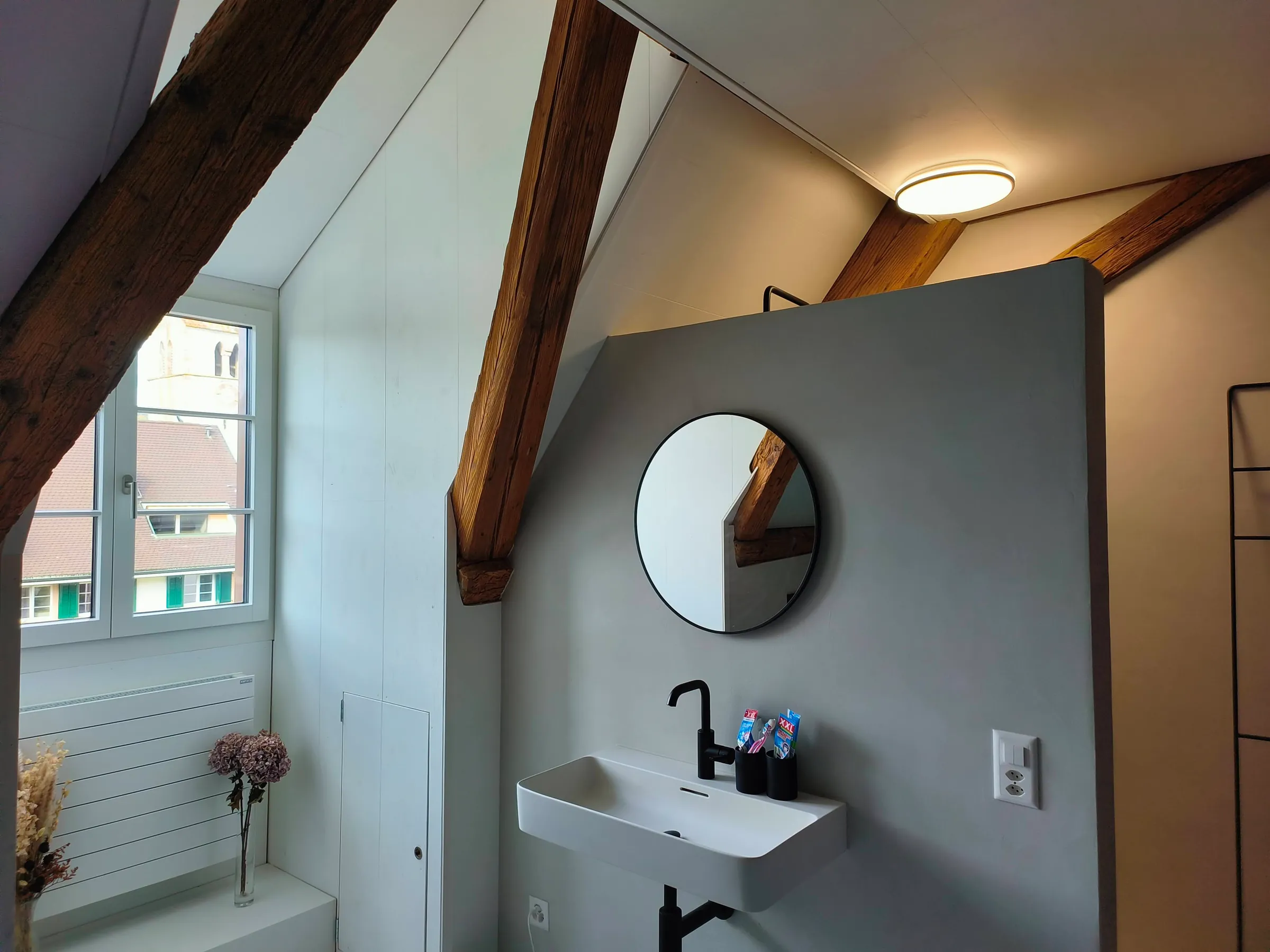 Badezimmer mit Waschtisch und rundem Spiegel unter altem Holzbalken, Fenster mit Blick auf den Kirchturm