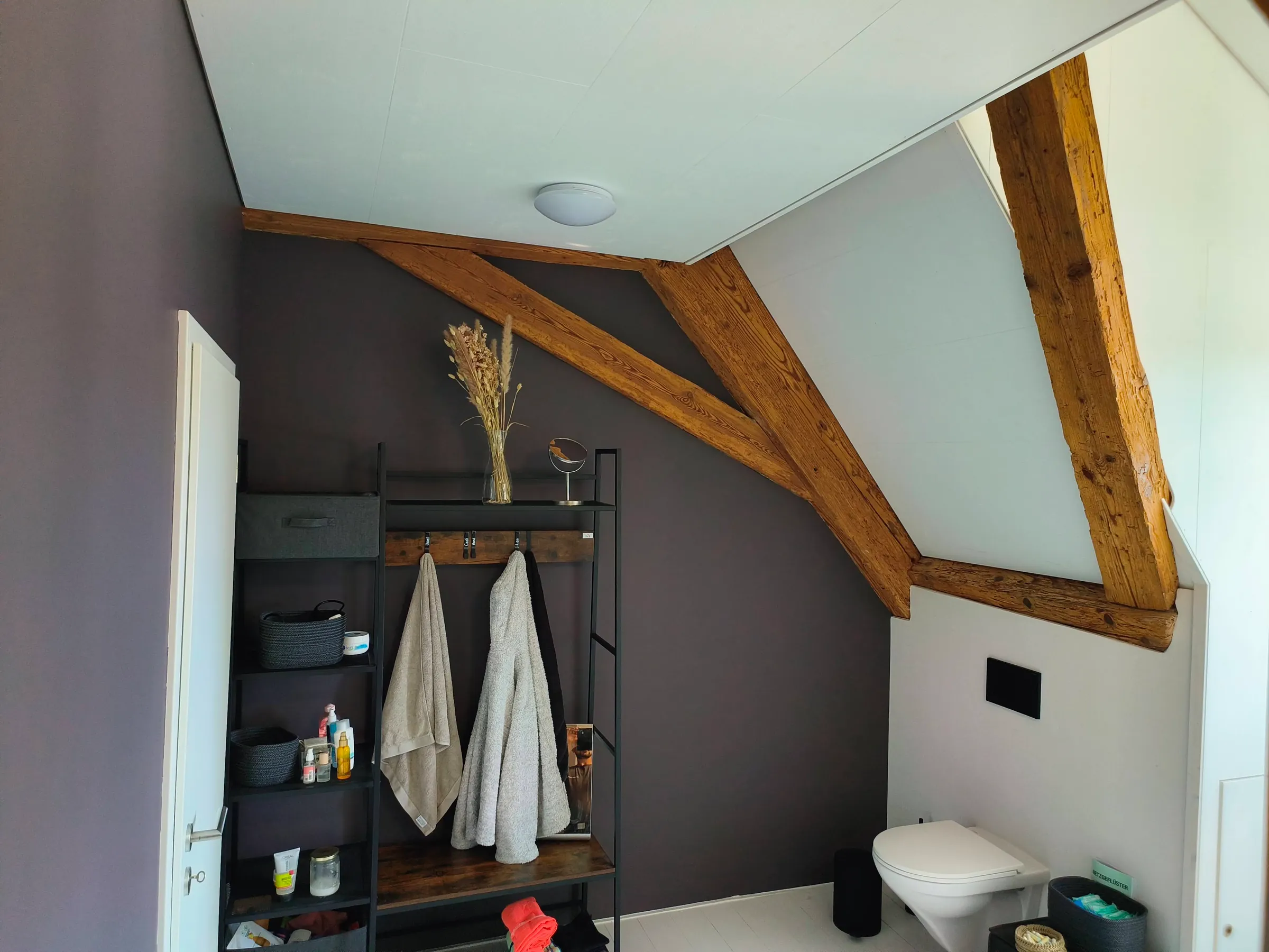 Badezimmer mit WC, dunkler Wand und alter Holzkonstruktion unter der Dachschräge