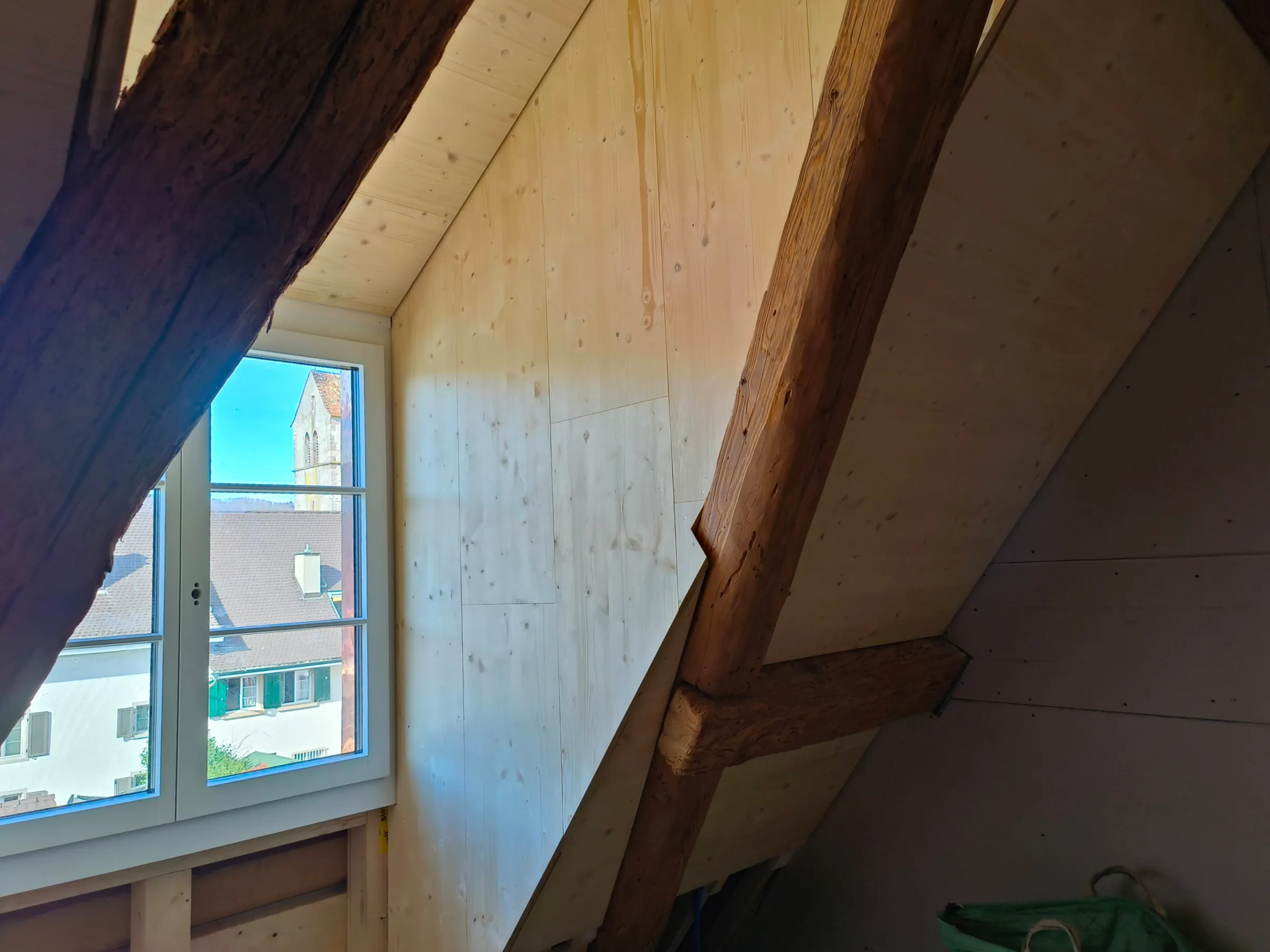Wand und Dachschräge mit hellen Dreischichtplatten verkleidet, alter Balken, Fenster mit Blick auf den Kirchturm