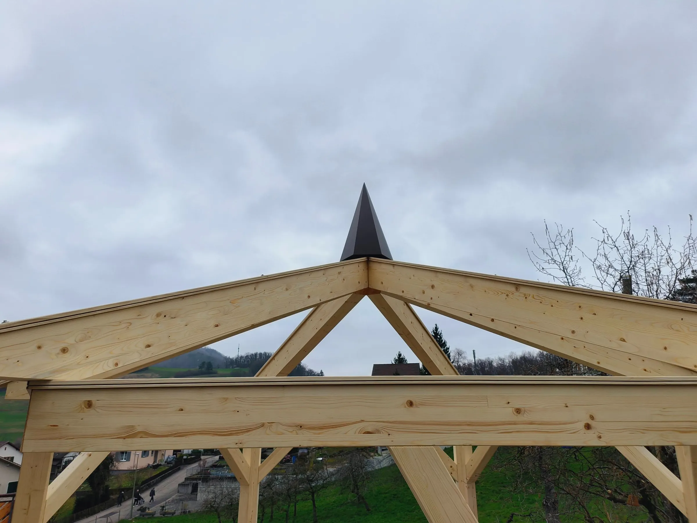 Sparren der Pergola laufen zur dekorativen Spitze aus Vollkernplatte zusammen, Dorf im Hintergrund
