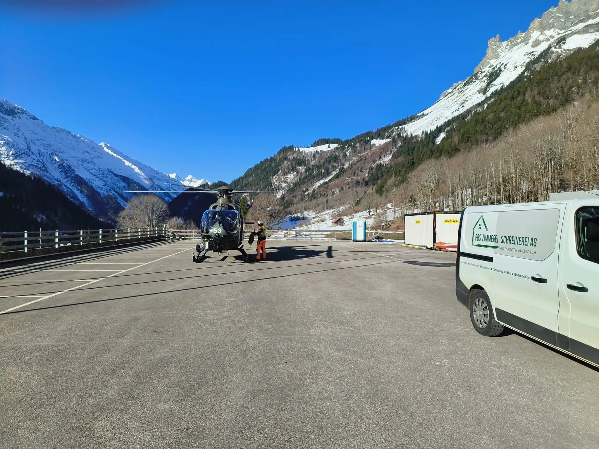 Militärhelikopter auf dem Parkplatz vor den Bergen, daneben Crew in Warnkleidung und der Firmentransporter